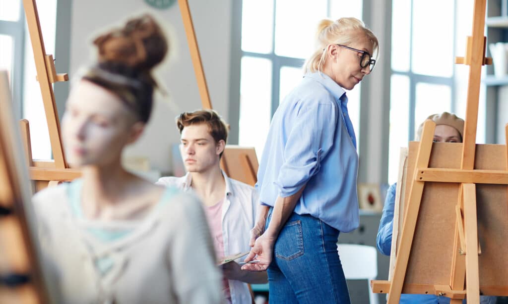 Female teacher walking along painting students at one of lessons in arts school
