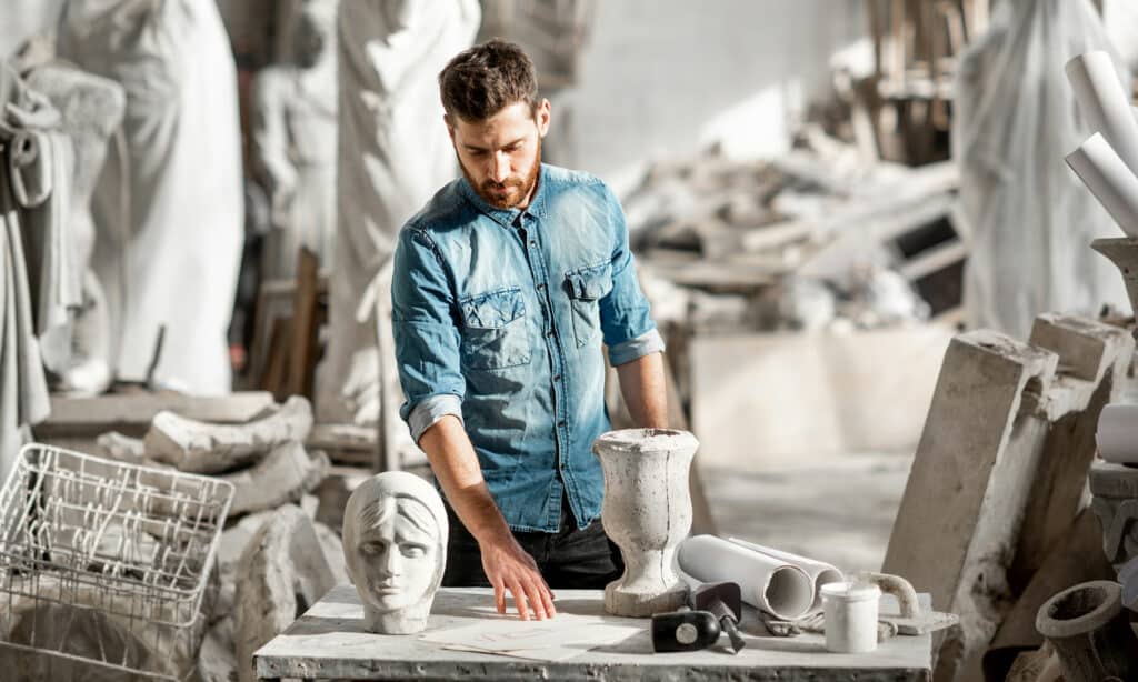 A portrait of a sculptor wearing a blue shirt working with stone sculpture on the table in his studio
